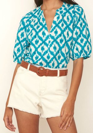 Vrouw draagt een teal en wit patroon blouse met een bruine gevlochten riem en witte high-waisted denim shorts.