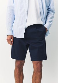 Granatowe szorty z miękkiego materiału z plisami z przodu, bocznymi kieszeniami i prostym krojem. Noszone z jasnoniebieską, paskowaną koszulą i białym t-shirtem.