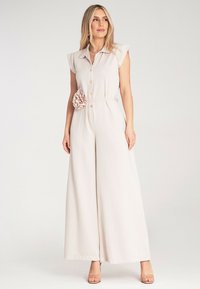 Jasnobeżowy kombinezon z krótkimi rękawkami, z przodu zapinany na guziki oraz z akcentem w postaci materiałowego kwiatu w talii. Rozszerzane nogawki, miękka tekstura, dopasowany krój.