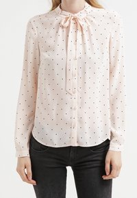 Blouse rose clair à pois noirs, manches longues, col à nouer en forme de nœud et devant boutonné. Tissu lisse avec un ourlet arrondi.