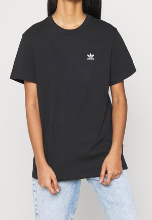 Nainen pukeutuneena mustaan Adidas-t-paitaan, jossa valkoinen logo rinnassa, ja vaaleansinisiin farkkuihin seisoo yksiväristä taustaa vasten.