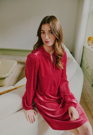 Une femme en robe rouge à motifs est assise sur un canapé blanc dans un salon lumineux.