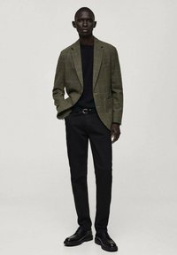 Blazer a quadri in verde tenue e marrone, abbinato a una camicia nera e pantaloni neri slim. Scarpe in pelle nera completano il look.