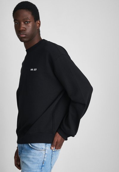 Sweatshirt noir fabriqué à partir d'un tissu doux et texturé. Il présente un col rond et un petit logo blanc "NN. 07" à l'avant. Associé à un denim bleu.