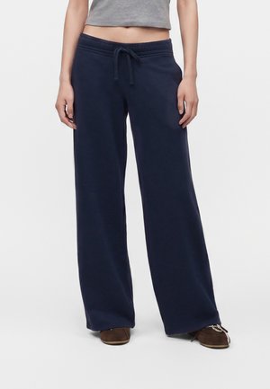 Personne portant un pantalon de survêtement bleu marine à jambes larges avec cordon à la taille et des chaussures marron, un haut gris court visible au-dessus du pantalon.