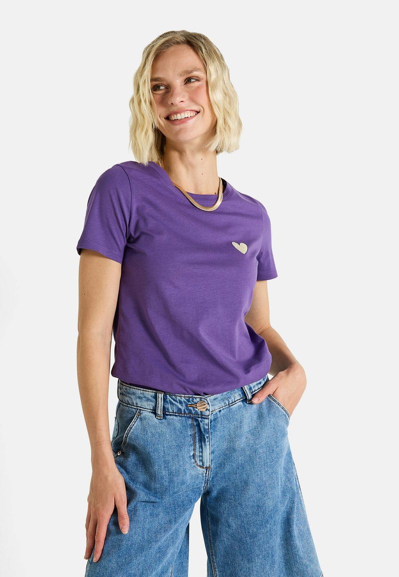 Camiseta de algodón morada de mangas cortas, con un pequeño detalle en forma de corazón dorado en el pecho. Combinada con jeans de corte holgado en azul claro.
