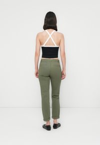 Kvinne som står med ryggen til iført en svart og hvit krysset crop top, olivengrønne bukser med oppbrett, og svarte flate sko på hvit bakgrunn.