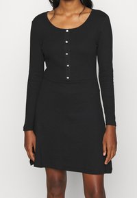 Svart långärmad klänning med rundad halsringning, med en knappslå och en figursydd överdel som fladdrar lätt vid fållen.