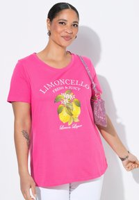 T-shirt en coton rose avec un col en V, arborant un graphique de citron et le texte "Limoncello Frais & Juteux." Accompagné d'un sac à main scintillant.
