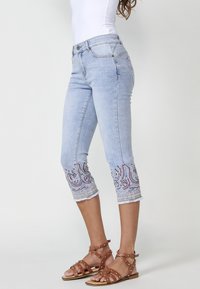 Light blue capri jeans in denim with embroidered detailing at the hem. Features a fitted design and a frayed edge. Paired with brown strap sandals.