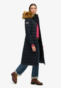 Marineblauwe gewatteerde lange jas met een met bont afgewerkte kap, ritssluiting aan de voorkant en logodetail op de mouw, gedragen over een roze trui en blauwe jeans.