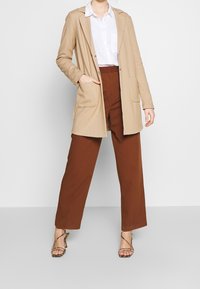 Beżowy długi blezer z klapami i przednimi kieszeniami, zestawiony z brązowymi spodniami o szerokich nogawkach. Noszony z sandałami na paski i białą koszulą z guzikami.