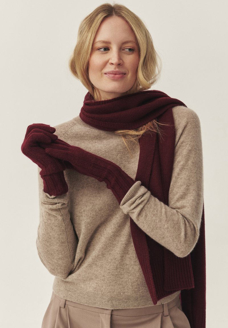 Fular burgund și mănuși fără degete asortate, din țesătură moale de tricot, purtate cu un pulover bej deschis, cu mâneci lungi și manșete striate.