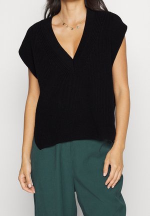 Kvinde iført sort ærmeløs strikket sweater med dyb V-hals og mørkegrønne bukser med høj talje, accessorized med en guld charms halskæde.