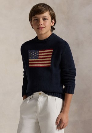 Garçon aux cheveux bruns portant un pull bleu marine avec un motif tricoté du drapeau américain et un pantalon blanc, debout devant un fond uni.