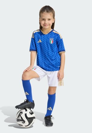 Mladá dívka v modrém dresu italského fotbalového týmu, bílých šortkách a modrých ponožkách stojí s jednou nohou na černobílém fotbalovém míči.