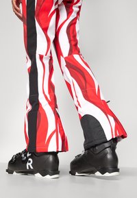 Schwarze Skischuhe mit drei weißen Schnallen, kombiniert mit rot-weißen Skihosen im Flammenmuster mit schwarzen Akzenten und mit Reißverschlussabschlüssen.