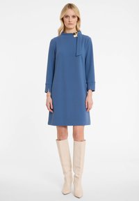 Robe bleue jusqu'aux genoux avec des manches longues, présentant un détail en nœud et un accent doré au niveau du cou. Portée avec des bottes montantes couleur crème.