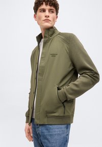 Férfi olívazöld Porsche BOSS dzsekiben, cipzáras zsebekkel és bordázott mandzsettákkal, kék farmerrel, oldalt nézve.