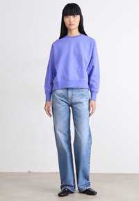 Joven mujer de pie, vestida con una sudadera lavanda, jeans rectos de lavado claro y zapatos negros, contra un fondo blanco liso.