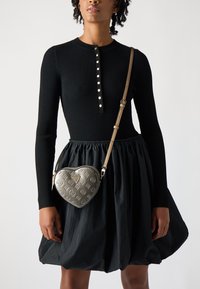 Top nero a coste con maniche lunghe e scollatura con bottoni, abbinato a una gonna nera voluminosa. Borsa a forma di cuore metallica con dettagli in rilievo.