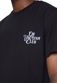 Czarna bawełniana koszulka z białym nadrukiem graficznym, na którym widnieje napis "ON VACATION CLUB" w stylizowanej czcionce na lewej stronie klatki piersiowej.