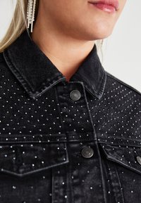 Sort denimjakke med klassisk krave, sølvfarvede nitter og to forlommer med metalknapper.