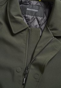 Veste vert foncé avec col, fermeture éclair partiellement ouverte, boutons-pression et doublure matelassée noire avec étiquette de la marque Matinique.