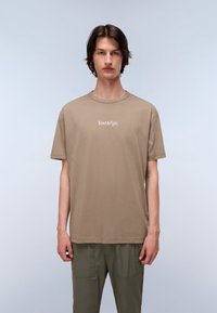 Brown cotton t-shirt featuring a crew neck, short sleeves, and a white logo print on the chest, paired with olive green pants.