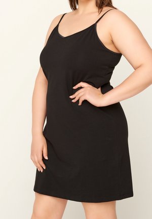 Vrouw die een mouwloze zwarte jurk met V-hals draagt, met de ene hand op haar heup en de andere hand langs haar zijde.