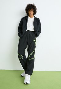 Mladić s kovrčavom kosom nosi crne Adidas trenirke s neonskim detaljima, crnu jaknu, bijelu majicu i bijele tenisice, stojeći na zelenom podu.