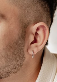 Orecchino a cerchio in argento indossato in un orecchio forato, caratterizzato da una superficie liscia e riflettente e da una forma circolare. Capelli corti e texturizzati visibili.