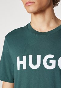 T-shirt en coton vert avec un col rond et un grand texte blanc en gras "HUGO" sur la poitrine. Texture lisse, manches courtes.