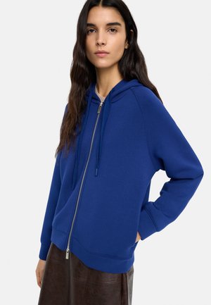 Młoda kobieta z długimi ciemnymi włosami w niebieskiej bluzie z zamkiem i ciemnobrązowej spódnicy, stojąca z jedną ręką w kieszeni.