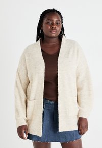 Crème-kleurige, gestructureerde gebreide cardigan met een open voorkant, met twee voorzakken, gedragen over een bruine top en een spijkerrok.