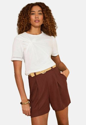 Žena s kučeravými vlasmi, oblečená v bielom krátkym rukávom tričku s detailom uzla a hnedých vysokých nohaviciach s béžovým opaskom.
