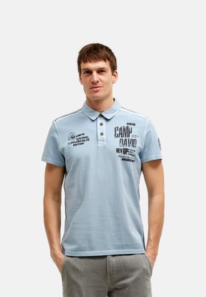 Mann trägt hellblaues Poloshirt mit schwarzen Textdrucken, steht mit Händen in den Taschen vor einfachem hellgrauem Hintergrund.