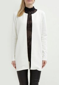 Vit texturerad kofta med rak skärning, öppen front och långa ärmar, lager-på-lager över en svart spets-topp och tillsammans med svarta byxor.