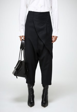 Zwarte broek met brede pijpen en een gedrapeerde voorkant, gecombineerd met een witte blouse. Het model houdt een kleine zwarte tas vast en draagt puntige, hoge laarzen.