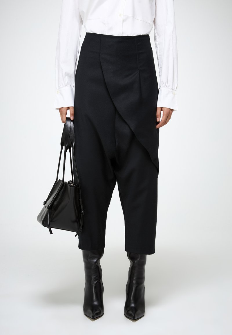 Zwarte broek met brede pijpen en een gedrapeerde voorkant, gecombineerd met een witte blouse. Het model houdt een kleine zwarte tas vast en draagt puntige, hoge laarzen.