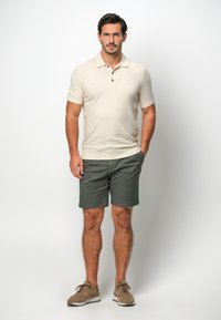 Beige Strick-Polohemd mit kurzen Ärmeln, Zwei-Knopf-Leiste, kombiniert mit dunkelgrünen Shorts und braunen Wildleder-Sneakern.