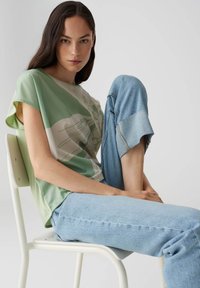 T-shirt astratto verde e beige con maniche corte, abbinato a jeans in denim azzurro chiaro con orli arrotolati, seduto su una sedia.