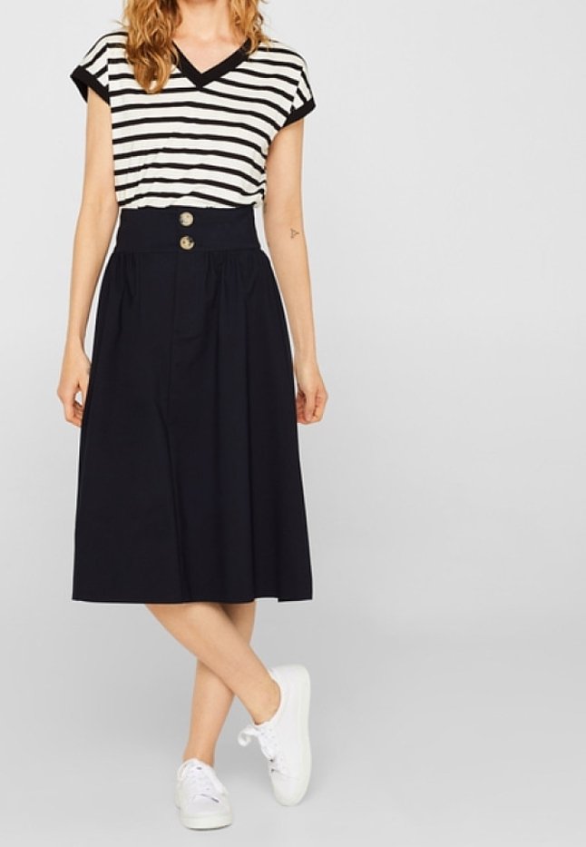 Top de rayas en blanco y negro de manga corta con escote en pico, combinado con una falda azul marino de cintura alta que presenta dos botones grandes y pliegues. Zapatillas blancas.