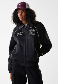 Juodas satinas bomber tipo švarkas su Niujorko "Yankees" logotipu, baltais akcentais ir elastinėmis rankovėmis. Modelis vilki bordo spalvos kepurę su tinkliniu nugaros skyriumi.