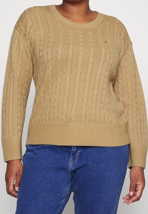 Osoba ubrana w beżowy sweter z grubym splotem i niebieskie dżinsy, stojąca z ramionami swobodnie opuszczonymi wzdłuż ciała na jednolitym tle.