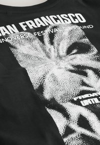 Schwarzes T-Shirt mit weißem, pixeligem Blumendesign und weißem Text mit der Aufschrift "San Francisco" sowie teilweise verdeckten Veranstaltungsdetails.