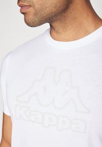 Camiseta de algodón blanca con un sutil logo en gris claro de "Kappa". La tela tiene una textura suave y un clásico escote redondeado.