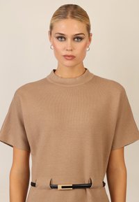 Beige Strickoberteil mit hohem Kragen und kurzen Ärmeln, versehen mit einem schwarzen Gürtel mit einer goldenen rechteckigen Schnalle. Glatte Textur.