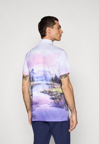 Muž stojaci bokom, oblečený v polo tričku s krátkym rukávom, na zadnej strane s potlačou pastelovej krajiny s horami a jazerom.