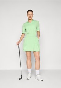 Polo de golf verde claro con cuello, combinado con una falda a juego con pliegues. La modelo sostiene un palo de golf y lleva zapatillas blancas.